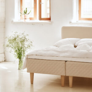 Matri Boxspringbett mit gestepptem, beigem Unterbau, weißen Kissen und Decke, neben einer Glasvase mit frischen Blumen.