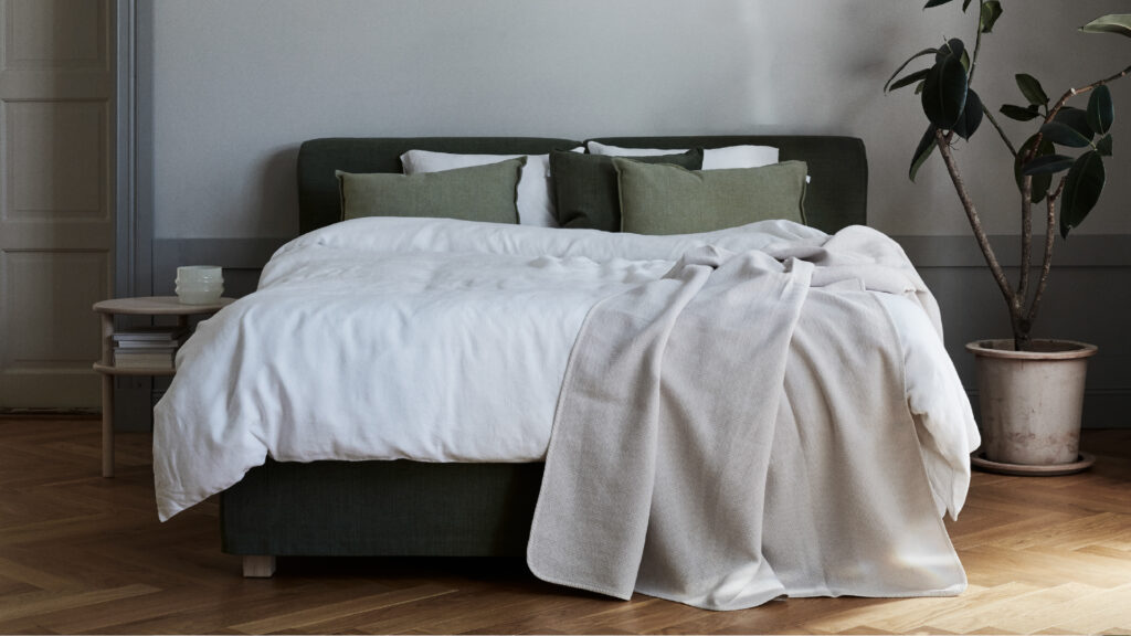 Matri Boxspringbett in Olivgrün, mit weißen und grünen Kissen, weißer Bettwäsche und beigem Überwurf, neben einem runden Holznachttisch und Pflanze.