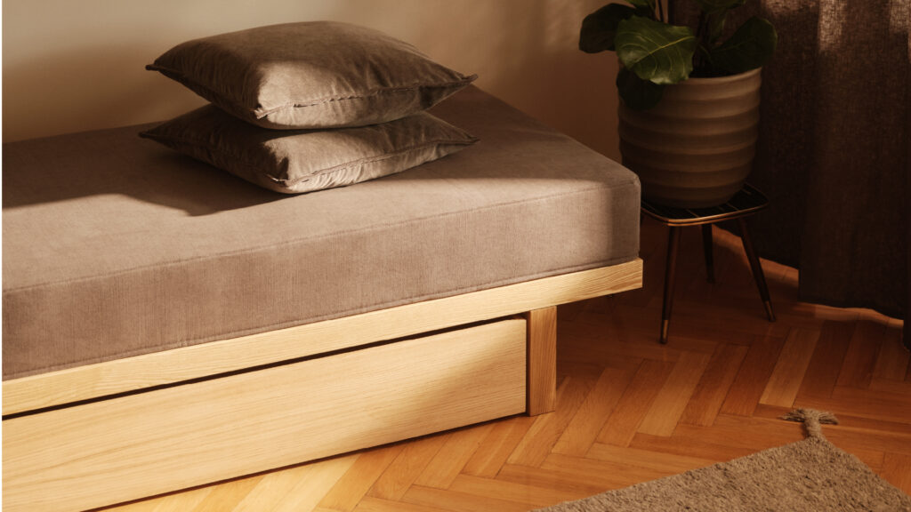 Matri Daybed aus hellem Holz mit integrierter Schublade, beigem Polster und zwei Kissen, neben einer großen Topfpflanze.