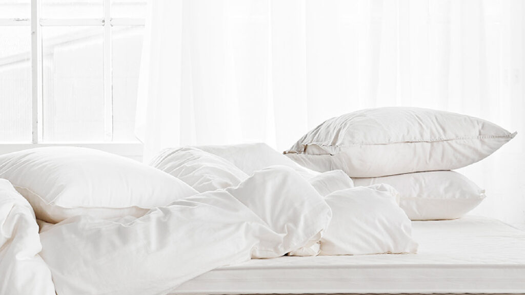 Bett mit weißer Matratze, darauf liegen weiße, weiche Matri Decken und Kissen vor einem hellen Fenster.