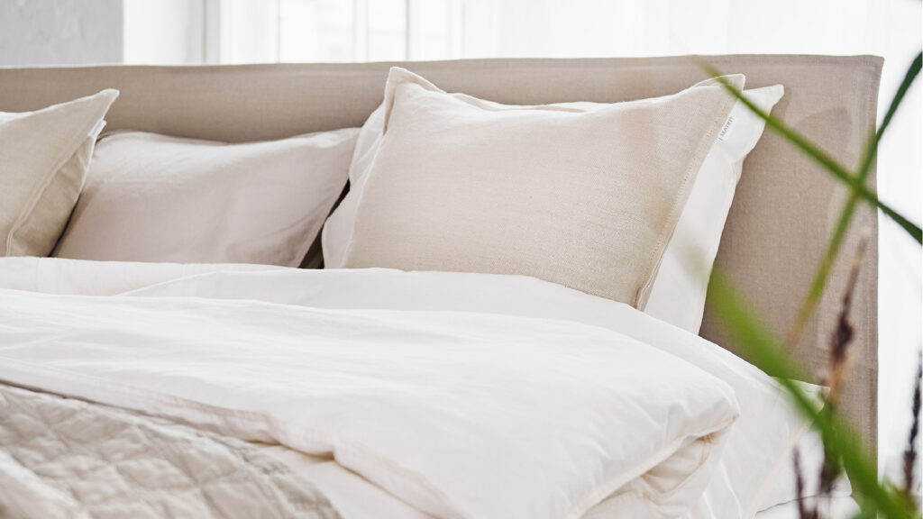 Detailaufnahme eines beigen Matri Kopfteil mit hellen Zierkissen und weißer Bettdecke.