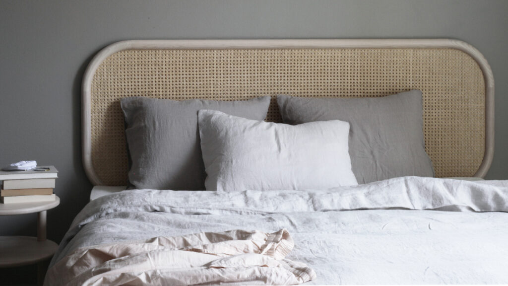 Matri Boxspringbett mit hellem Rattan-Kopfteil, Leinenbettwäsche in Hellgrau und Weiß sowie einem kleinen Nachttisch.