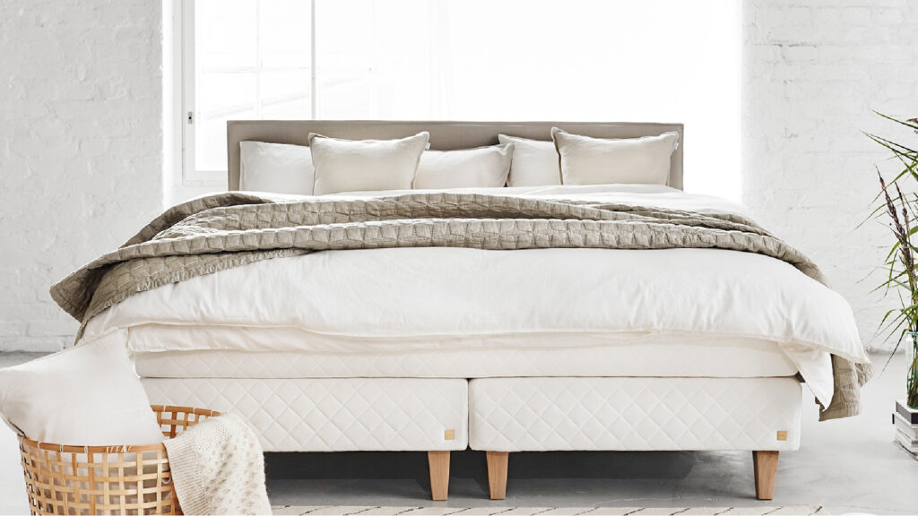 Großes, helles Boxspringbett mit gestepptem Sockel, schlichtem Kopfteil, weißer Bettwäsche und grob gestricktem beigem Überwurf im Loft-Schlafzimmer.